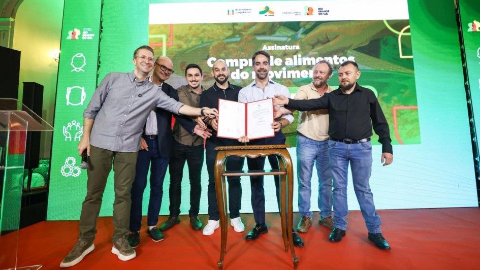 Foto mostra governador Eduardo Leite com documento em m&atilde;os, tendo pessoas ao seu lado, no ato do Pal&aacute;cio Piratini