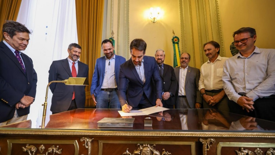 Foto de Leite assinando, de p&eacute;, um documento em uma mesa. Ao seu redor h&aacute; sete homens acompanhando a cena em ambiente formal.
