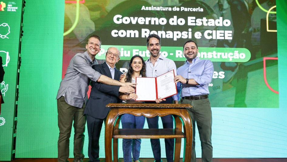 Uma fotografia em um evento interno, com fundo verde-claro e um letreiro grande na parte superior. Quatro homens e uma mulher est&atilde;o ao redor de uma pequena mesa de madeira escura com tampo oval, no centro da imagem.

Eles est&atilde;o em p&eacute;, sorrindo e estendendo as m&atilde;os para um documento que est&aacute; sendo segurado pela mulher. A mulher, vestindo uma cal&ccedil;a jeans escura e uma blusa azul-claro, est&aacute; no centro da mesa, segurando o documento vermelho (uma pasta ou certificado). O homem &agrave; sua direita (esquerda na foto), mais velho, de terno cinza, apoia a m&atilde;o sobre a dela. O homem &agrave; esquerda (direita na foto), de camisa clara com as mangas arrega&ccedil;adas, tamb&eacute;m apoia a m&atilde;o sobre a da mulher. Outros dois homens, um de camisa cinza (na extrema esquerda) e outro de camisa azul-escura (na extrema direita), est&atilde;o olhando para a c&acirc;mera e sorrindo, com as m&atilde;os espalmadas ou levemente levantadas, pr&oacute;ximos ao grupo central.

O letreiro acima diz: "Assinatura de Parceria - Governo do Estado com Renapsi e CIEE" e logo abaixo, em letras maiores: "Partiu Futuro - Reconstru&ccedil;&atilde;o".

A ilumina&ccedil;&atilde;o &eacute; clara e focada no grupo de pessoas.