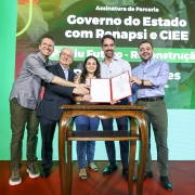 Uma fotografia em um evento interno, com fundo verde-claro e um letreiro grande na parte superior. Quatro homens e uma mulher est&atilde;o ao redor de uma pequena mesa de madeira escura com tampo oval, no centro da imagem.

Eles est&atilde;o em p&eacute;, sorrindo e estendendo as m&atilde;os para um documento que est&aacute; sendo segurado pela mulher. A mulher, vestindo uma cal&ccedil;a jeans escura e uma blusa azul-claro, est&aacute; no centro da mesa, segurando o documento vermelho (uma pasta ou certificado). O homem &agrave; sua direita (esquerda na foto), mais velho, de terno cinza, apoia a m&atilde;o sobre a dela. O homem &agrave; esquerda (direita na foto), de camisa clara com as mangas arrega&ccedil;adas, tamb&eacute;m apoia a m&atilde;o sobre a da mulher. Outros dois homens, um de camisa cinza (na extrema esquerda) e outro de camisa azul-escura (na extrema direita), est&atilde;o olhando para a c&acirc;mera e sorrindo, com as m&atilde;os espalmadas ou levemente levantadas, pr&oacute;ximos ao grupo central.

O letreiro acima diz: "Assinatura de Parceria - Governo do Estado com Renapsi e CIEE" e logo abaixo, em letras maiores: "Partiu Futuro - Reconstru&ccedil;&atilde;o".

A ilumina&ccedil;&atilde;o &eacute; clara e focada no grupo de pessoas.