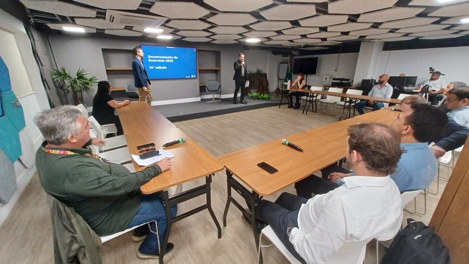 Foto mostra uma sala ampla, com um palestrante falando ao microfone ao fundo, e ao redor, sentados atr&aacute;s de mesas no formato U, est&atilde;o outros homens que acompanham a apresenta&ccedil;&atilde;o.