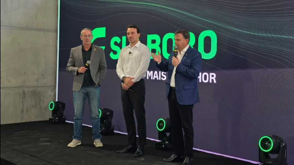 Uma fotografia de tr&ecirc;s homens em um palco durante um evento de inaugura&ccedil;&atilde;o em um ambiente interno.

O palco &eacute; simples, com o ch&atilde;o escuro e algumas luzes de LED verdes no ch&atilde;o, dispostas em formato de anel. O fundo &eacute; um grande painel digital ou tela escura com padr&otilde;es de linhas verdes vibrantes e o texto grande "SU BORO" (com a s&iacute;laba "SU" em verde) seguido por "MAIS..." e a palavra "HOR" no canto inferior direito, sugerindo uma frase como "MAIS MELHOR".

Tr&ecirc;s homens est&atilde;o em p&eacute;, lado a lado, voltados para a frente:

Homem &agrave; esquerda: Veste jeans, um su&eacute;ter escuro e um blazer cinza-claro. Ele segura um microfone com a m&atilde;o direita, mas n&atilde;o est&aacute; falando, e tem um sorriso contido.

Homem no centro: Veste uma camisa social branca de manga comprida e cal&ccedil;a escura. Ele est&aacute; com as m&atilde;os cruzadas &agrave; frente do corpo e olha para o homem que est&aacute; falando.

Homem &agrave; direita: Veste uma camisa clara, cal&ccedil;a escura e um blazer azul. Ele est&aacute; falando no microfone que segura com a m&atilde;o esquerda, gesticulando com a m&atilde;o direita aberta.

A ilumina&ccedil;&atilde;o &eacute; focada nos participantes e na tela de fundo. O ambiente parece ser um galp&atilde;o industrial ou um local amplo, com uma parede cinza-concreto vis&iacute;vel na parte superior.