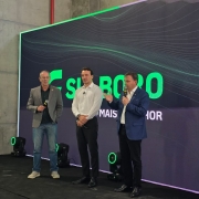 Uma fotografia de tr&ecirc;s homens em um palco durante um evento de inaugura&ccedil;&atilde;o em um ambiente interno.

O palco &eacute; simples, com o ch&atilde;o escuro e algumas luzes de LED verdes no ch&atilde;o, dispostas em formato de anel. O fundo &eacute; um grande painel digital ou tela escura com padr&otilde;es de linhas verdes vibrantes e o texto grande "SU BORO" (com a s&iacute;laba "SU" em verde) seguido por "MAIS..." e a palavra "HOR" no canto inferior direito, sugerindo uma frase como "MAIS MELHOR".

Tr&ecirc;s homens est&atilde;o em p&eacute;, lado a lado, voltados para a frente:

Homem &agrave; esquerda: Veste jeans, um su&eacute;ter escuro e um blazer cinza-claro. Ele segura um microfone com a m&atilde;o direita, mas n&atilde;o est&aacute; falando, e tem um sorriso contido.

Homem no centro: Veste uma camisa social branca de manga comprida e cal&ccedil;a escura. Ele est&aacute; com as m&atilde;os cruzadas &agrave; frente do corpo e olha para o homem que est&aacute; falando.

Homem &agrave; direita: Veste uma camisa clara, cal&ccedil;a escura e um blazer azul. Ele est&aacute; falando no microfone que segura com a m&atilde;o esquerda, gesticulando com a m&atilde;o direita aberta.

A ilumina&ccedil;&atilde;o &eacute; focada nos participantes e na tela de fundo. O ambiente parece ser um galp&atilde;o industrial ou um local amplo, com uma parede cinza-concreto vis&iacute;vel na parte superior.