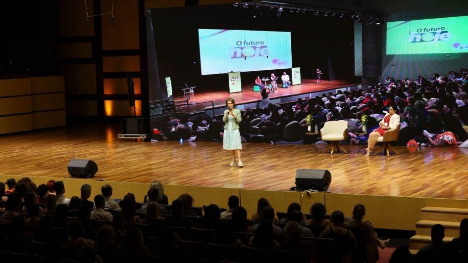 Secret&aacute;ria Raquel Teixeira est&aacute; num palco de piso de madeira polida. Ela fala em p&eacute; para uma plateia. Atr&aacute;s dela, um tel&atilde;o gigante reproduz a foto de outro evento, tamb&eacute;m em um audit&oacute;rio.