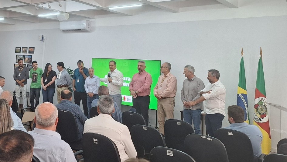 Foto mostra secret&aacute;rio falando em p&eacute; ao microfone, com prefeitos, lado a lado, enfileirados, acompanhando o discurso. Eles est&atilde;o diante de uma plateia sentada. Atr&aacute;s do palco h&aacute; um tel&atilde;o e bandeiras. 