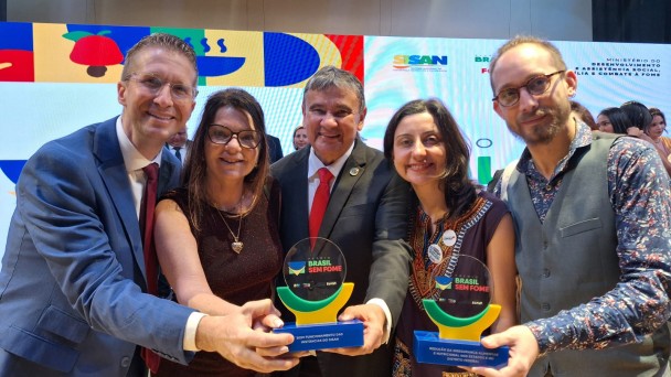
Esta &eacute; uma fotografia em plano m&eacute;dio e close-up de cinco pessoas (tr&ecirc;s homens e duas mulheres) sorrindo e posando enquanto seguram dois trof&eacute;us em uma cerim&ocirc;nia de premia&ccedil;&atilde;o.

As Pessoas (da esquerda para a direita):

Homem 1: Veste terno azul, camisa branca e gravata bord&ocirc;. Ele usa &oacute;culos de grau e sorri abertamente.

Mulher 1: Tem cabelos castanhos longos, usa &oacute;culos de grau e um vestido texturizado em tom marrom escuro ou vinho. Ela usa um colar com pingente de cora&ccedil;&atilde;o dourado.

Homem 2 (ao centro): Um homem mais velho, vestindo terno preto, camisa branca e gravata vermelha. Ele est&aacute; posicionado no centro, com os bra&ccedil;os estendidos para segurar os dois trof&eacute;us junto com os colegas.

Mulher 2: Tem cabelos escuros e veste uma roupa escura com um detalhe bordado colorido (estilo &eacute;tnico ou tribal) no decote. Ela sorri diretamente para a c&acirc;mera.

Homem 3 (&agrave; direita): Veste uma camisa estampada azul com flores pequenas por baixo de um colete cinza. Ele tem barba curta, usa &oacute;culos e possui um pequeno brinco na orelha esquerda.

Os Trof&eacute;us: Eles seguram dois trof&eacute;us id&ecirc;nticos de design moderno.

A base &eacute; um ret&acirc;ngulo azul escuro com texto informativo em branco.

Sobre a base, h&aacute; um suporte em formato de semic&iacute;rculo (meia-lua) nas cores verde e amarelo.

A parte superior &eacute; um disco de acr&iacute;lico transparente onde se l&ecirc;: "PR&Ecirc;MIO BRASIL SEM FOME" em letras coloridas.

O trof&eacute;u da esquerda traz o texto na base: "BOM FUNCIONAMENTO DAS INST&Acirc;NCIAS DO SISAN".

O trof&eacute;u da direita traz: "REDU&Ccedil;&Atilde;O DA INSEGURAN&Ccedil;A ALIMENTAR E NUTRICIONAL NOS ESTADOS E NO DISTRITO FEDERAL".

O Fundo: Atr&aacute;s do grupo, h&aacute; uma grande tela de LED com gr&aacute;ficos vibrantes e abstratos em amarelo, vermelho, azul e verde. No canto superior direito da tela, &eacute; vis&iacute;vel o logotipo do SISAN e do Minist&eacute;rio do Desenvolvimento e Assist&ecirc;ncia Social, Fam&iacute;lia e Combate &agrave; Fome.

A cena &eacute; de celebra&ccedil;&atilde;o e vit&oacute;ria, com ilumina&ccedil;&atilde;o clara e foco centralizado no grupo e nos pr&ecirc;mios.