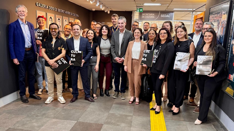 
Esta &eacute; uma fotografia em plano geral de um grupo numeroso de pessoas (cerca de 25 a 30 indiv&iacute;duos) posando para uma foto oficial em um ambiente interno institucional.

O Grupo:

Composto por homens e mulheres de diversas idades, vestidos predominantemente com trajes casuais e profissionais (blazers, camisas, blusas e cal&ccedil;as sociais).

Est&atilde;o distribu&iacute;dos de p&eacute;, ocupando o centro e as laterais do quadro.

Pelo menos cinco pessoas, distribu&iacute;das pelo grupo, seguram &agrave; frente do corpo uma pasta ou livreto preto com a palavra "CULTURA" escrita verticalmente em letras brancas grandes e em negrito.

A maioria das pessoas est&aacute; sorrindo, voltada para a c&acirc;mera.

O Ambiente:

&Agrave; esquerda: H&aacute; uma parede marrom escura com a inscri&ccedil;&atilde;o em letras met&aacute;licas: "GALERIA DOS EX-SECRET&Aacute;RIOS DA CULTURA". Abaixo do texto, h&aacute; v&aacute;rias molduras com retratos fotogr&aacute;ficos de antigos gestores alinhados em duas fileiras.

Ao fundo: No centro, acima de um corredor, h&aacute; uma placa de sinaliza&ccedil;&atilde;o cinza que indica: "SECRETARIA DA CULTURA SEDAC". A ilumina&ccedil;&atilde;o do teto &eacute; feita por trilhos com spots direcionais brancos.

&Agrave; direita: Parte de uma parede exibe um cartaz ou obra de arte emoldurada com elementos gr&aacute;ficos coloridos.

O ch&atilde;o: Revestido com grandes lajetas de cer&acirc;mica cinza. No lado direito, destaca-se uma faixa de piso t&aacute;til de alerta na cor amarela, que atravessa verticalmente parte do cen&aacute;rio.

A imagem registra um encontro ou evento oficial na sede da Secretaria de Estado da Cultura (Sedac) do Rio Grande do Sul.