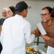 Foto mostra alunos do curso recebendo os certificados. Eles est&atilde;o de costas para a foto e de frente para os respons&aacute;veis pelo curso que entregam os certificados. Em destaque, um deles, de bon&eacute; preto e camisa branca, &eacute; cumprimentado por um homem de &oacute;culos que o sa&uacute;da com um aperto de m&atilde;o e est&aacute; muito sorridente.