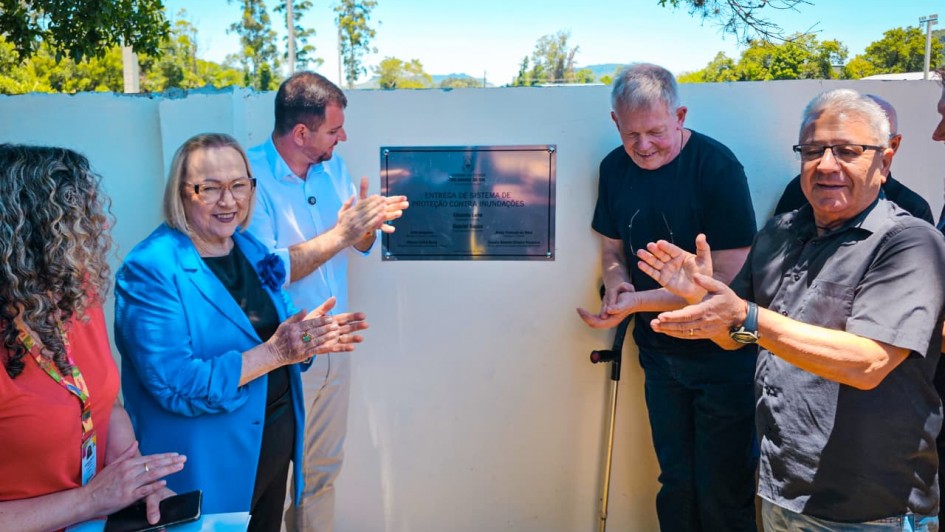 Foto ao ar livre de cinco pessoas aplaudindo ao redor da placa de inaugura&ccedil;&atilde;o, pregada num muro branco baixo.