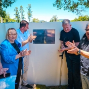 Foto ao ar livre de cinco pessoas aplaudindo ao redor da placa de inaugura&ccedil;&atilde;o, pregada num muro branco baixo.