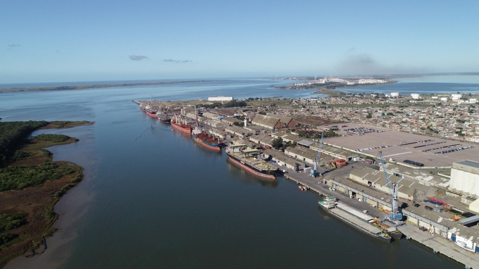 Imagem a&eacute;rea de um porto p&uacute;blico &agrave;s margens de um rio largo e calmo. V&aacute;rios navios de carga de grande porte est&atilde;o atracados em sequ&ecirc;ncia ao longo dos cais. &Agrave; direita, h&aacute; extensos armaz&eacute;ns portu&aacute;rios, guindastes e &aacute;reas pavimentadas para opera&ccedil;&atilde;o log&iacute;stica. &Agrave; esquerda do rio, aparecem &aacute;reas de vegeta&ccedil;&atilde;o baixa e ilhas fluviais. Ao fundo, observa-se a foz e a continuidade do curso d&rsquo;&aacute;gua em dire&ccedil;&atilde;o ao horizonte, sob c&eacute;u azul claro.