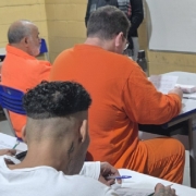 Foto mostra quatro apenados sentados em carteiras de escola. Eles est&atilde;o de costas para a foto. Tr&ecirc;s deles vestem uniformes laranjas.