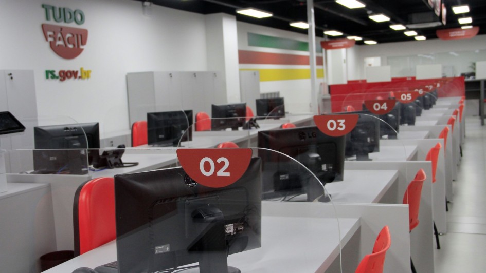 Imagem mostra uma fileira de guich&ecirc;s do Tudo F&aacute;cil de Passo Fundo, com computadores sobre as mesas cinzas, cadeiras laranjas e identifica&ccedil;&atilde;o com os n&uacute;meros dos guich&ecirc;s em placas vermelhas com n&uacute;meros brancos. Ao fundo parede branca com s&iacute;mbolo do Tudo F&aacute;cil.