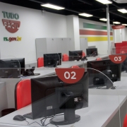 Imagem mostra uma fileira de guich&ecirc;s do Tudo F&aacute;cil de Passo Fundo, com computadores sobre as mesas cinzas, cadeiras laranjas e identifica&ccedil;&atilde;o com os n&uacute;meros dos guich&ecirc;s em placas vermelhas com n&uacute;meros brancos. Ao fundo parede branca com s&iacute;mbolo do Tudo F&aacute;cil.