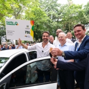 Foto ao ar livre posada, de Leite segurando uma grande r&eacute;plica de um cart&atilde;o cidad&atilde;o nas m&atilde;os, enquanto sorri. Ele est&aacute; ao lado de um dos ve&iacute;culos entregues. H&aacute; quatro homens junto a ele no primeiro plano, e v&aacute;rias ao fundo, num ambiente arborizado.
