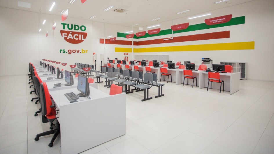 Foto mostra imagem da sala da ag&ecirc;ncia, com muitos guich&ecirc;s na cor cinza claro e cadeiras laranjas e sobre eles computadores. Ao fundo, a logomarca do Tudo F&aacute;cil e listas amarelas vermelhas e verdes decorando as paredes. 