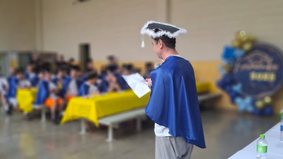 Foto de um homem com uma beca azul discursando ao microfone, meio de costas para a c&acirc;mera, de modo que n&atilde;o &eacute; poss&iacute;vel ver seu rosto. Ao fundo, a imagem aparece emba&ccedil;ada, mas &eacute; poss&iacute;vel ver v&aacute;rias pessoas de beca assistindo.