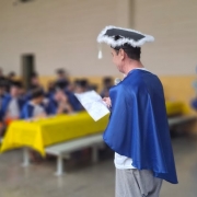 Foto de um homem com uma beca azul discursando ao microfone, meio de costas para a c&acirc;mera, de modo que n&atilde;o &eacute; poss&iacute;vel ver seu rosto. Ao fundo, a imagem aparece emba&ccedil;ada, mas &eacute; poss&iacute;vel ver v&aacute;rias pessoas de beca assistindo.