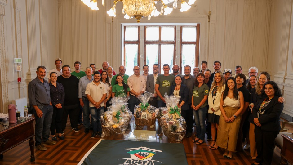 Foto posada de mais de 30 pessoas, em p&eacute; e de frente para a c&acirc;mera, num ambiente fechado e solene. &Agrave; frente, h&aacute; tr&ecirc;s cestas embrulhadas com objetos que n&atilde;o se consegue identificar.