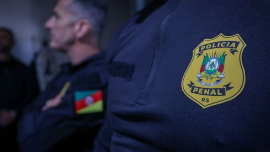 Foto do detalhe do uniforme, de cor escura, de um policial penal. O destaque &eacute; o escudo, de cor amarelo, no qual est&aacute; escrita Pol&iacute;cia Penal RS