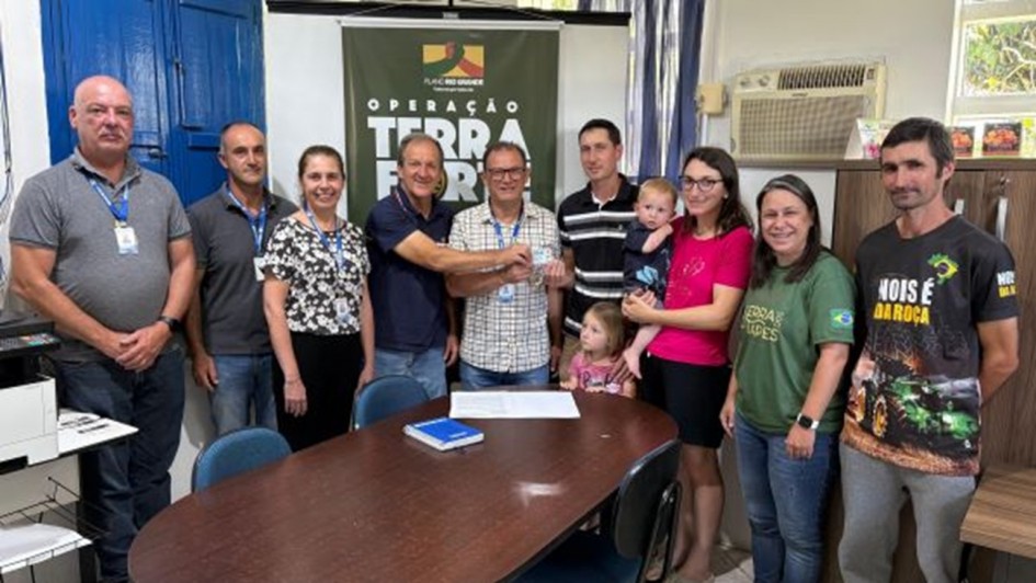 Uma fotografia interna mostra um grupo de onze pessoas &mdash; sete adultos, duas crian&ccedil;as e um beb&ecirc; &mdash; reunidas em torno de uma mesa oval de madeira escura em um escrit&oacute;rio. Ao fundo, um banner verde-oliva exibe o logotipo do "Plano Rio Grande" e as palavras "OPERA&Ccedil;&Atilde;O TERRA FORTE" em letras brancas grandes.

No centro da imagem, dois homens seguram juntos um pequeno cart&atilde;o branco, simbolizando um ato de entrega. O homem &agrave; esquerda usa uma camisa polo azul-marinho, e o homem &agrave; direita veste uma camisa de mangas curtas quadriculada. &Agrave; direita deles, um homem jovem com camisa listrada observa o ato, enquanto uma mulher de camiseta rosa segura um beb&ecirc; no colo. Uma menina pequena est&aacute; posicionada &agrave; frente deles, pr&oacute;xima &agrave; mesa.

Do lado esquerdo da mesa, tr&ecirc;s adultos (dois homens de cinza e uma mulher com blusa estampada) sorriem para a c&acirc;mera. No lado direito, uma mulher com camiseta verde da "Terra de Todos" e um homem com uma camiseta preta estampada com a frase "Nois &eacute; da Ro&ccedil;a" completam o grupo.

O ambiente &eacute; simples, com paredes brancas, uma janela com moldura azul &agrave; esquerda, um aparelho de ar-condicionado de janela &agrave; direita e equipamentos de escrit&oacute;rio, como uma impressora, vis&iacute;veis nas extremidades. Sobre a mesa, h&aacute; um caderno azul e algumas folhas de papel.