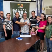 Uma fotografia interna mostra um grupo de onze pessoas &mdash; sete adultos, duas crian&ccedil;as e um beb&ecirc; &mdash; reunidas em torno de uma mesa oval de madeira escura em um escrit&oacute;rio. Ao fundo, um banner verde-oliva exibe o logotipo do "Plano Rio Grande" e as palavras "OPERA&Ccedil;&Atilde;O TERRA FORTE" em letras brancas grandes.

No centro da imagem, dois homens seguram juntos um pequeno cart&atilde;o branco, simbolizando um ato de entrega. O homem &agrave; esquerda usa uma camisa polo azul-marinho, e o homem &agrave; direita veste uma camisa de mangas curtas quadriculada. &Agrave; direita deles, um homem jovem com camisa listrada observa o ato, enquanto uma mulher de camiseta rosa segura um beb&ecirc; no colo. Uma menina pequena est&aacute; posicionada &agrave; frente deles, pr&oacute;xima &agrave; mesa.

Do lado esquerdo da mesa, tr&ecirc;s adultos (dois homens de cinza e uma mulher com blusa estampada) sorriem para a c&acirc;mera. No lado direito, uma mulher com camiseta verde da "Terra de Todos" e um homem com uma camiseta preta estampada com a frase "Nois &eacute; da Ro&ccedil;a" completam o grupo.

O ambiente &eacute; simples, com paredes brancas, uma janela com moldura azul &agrave; esquerda, um aparelho de ar-condicionado de janela &agrave; direita e equipamentos de escrit&oacute;rio, como uma impressora, vis&iacute;veis nas extremidades. Sobre a mesa, h&aacute; um caderno azul e algumas folhas de papel.