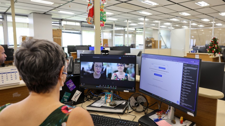 A fotografia mostra uma mulher de costas, em primeiro plano, sentada &agrave; sua mesa em um escrit&oacute;rio de plano aberto. Ela tem cabelos curtos e grisalhos e usa uma blusa colorida com estampa floral.

&Agrave; sua frente, h&aacute; dois monitores de computador:

Monitor da esquerda: Exibe uma videochamada em andamento com duas janelas de v&iacute;deo. Uma mostra a pr&oacute;pria mulher que est&aacute; de costas e a outra mostra uma mulher mais jovem usando &oacute;culos e fones de ouvido.

Monitor da direita: Mostra uma tela de sistema ou documento com texto em portugu&ecirc;s e uma lista de arquivos baixados na lateral direita.

A mesa de trabalho est&aacute; organizada com diversos itens, incluindo um teclado preto, um mouse, um pequeno ventilador USB, um calend&aacute;rio da Caixa Econ&ocirc;mica Federal e alguns objetos decorativos, como uma pequena imagem de um gato.

O ambiente de escrit&oacute;rio ao fundo &eacute; amplo, com divis&oacute;rias baixas, teto de placas brancas e ilumina&ccedil;&atilde;o fluorescente. &Eacute; poss&iacute;vel ver outras mesas de trabalho e um pequeno enfeite de Natal (uma &aacute;rvore dourada) em uma mesa ao fundo &agrave; direita, sugerindo que a foto foi tirada pr&oacute;ximo ao final do ano.

O t&iacute;tulo do arquivo indica que a cena retrata o momento da assinatura de uma escritura p&uacute;blica de doa&ccedil;&atilde;o de um im&oacute;vel do Estado para o campus do IFFar (Instituto Federal Farroupilha) em Santiago, realizada de forma digital/remota.