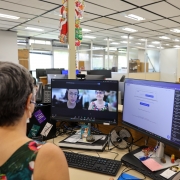 A fotografia mostra uma mulher de costas, em primeiro plano, sentada &agrave; sua mesa em um escrit&oacute;rio de plano aberto. Ela tem cabelos curtos e grisalhos e usa uma blusa colorida com estampa floral.

&Agrave; sua frente, h&aacute; dois monitores de computador:

Monitor da esquerda: Exibe uma videochamada em andamento com duas janelas de v&iacute;deo. Uma mostra a pr&oacute;pria mulher que est&aacute; de costas e a outra mostra uma mulher mais jovem usando &oacute;culos e fones de ouvido.

Monitor da direita: Mostra uma tela de sistema ou documento com texto em portugu&ecirc;s e uma lista de arquivos baixados na lateral direita.

A mesa de trabalho est&aacute; organizada com diversos itens, incluindo um teclado preto, um mouse, um pequeno ventilador USB, um calend&aacute;rio da Caixa Econ&ocirc;mica Federal e alguns objetos decorativos, como uma pequena imagem de um gato.

O ambiente de escrit&oacute;rio ao fundo &eacute; amplo, com divis&oacute;rias baixas, teto de placas brancas e ilumina&ccedil;&atilde;o fluorescente. &Eacute; poss&iacute;vel ver outras mesas de trabalho e um pequeno enfeite de Natal (uma &aacute;rvore dourada) em uma mesa ao fundo &agrave; direita, sugerindo que a foto foi tirada pr&oacute;ximo ao final do ano.

O t&iacute;tulo do arquivo indica que a cena retrata o momento da assinatura de uma escritura p&uacute;blica de doa&ccedil;&atilde;o de um im&oacute;vel do Estado para o campus do IFFar (Instituto Federal Farroupilha) em Santiago, realizada de forma digital/remota.