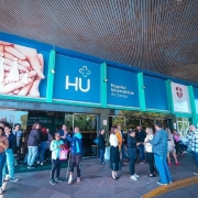 Esta &eacute; uma descri&ccedil;&atilde;o da imagem para fins de acessibilidade:

A fotografia mostra a fachada e a entrada principal do Hospital Universit&aacute;rio de Canoas (HU) em um dia claro e ensolarado.

Detalhes da Fachada
O pr&eacute;dio apresenta uma estrutura moderna com pain&eacute;is em tons de azul vibrante e verde.

&Agrave; esquerda, h&aacute; um grande painel art&iacute;stico mostrando m&atilde;os que protegem a silhueta de uma fam&iacute;lia (pai, m&atilde;e e duas crian&ccedil;as).

Ao centro, destaca-se o logotipo do hospital: uma cruz estilizada acima das letras grandes "HU", seguidas pelo texto "Hospital Universit&aacute;rio de Canoas".

&Agrave; direita, h&aacute; outros logotipos menores, incluindo o da associa&ccedil;&atilde;o que faz a gest&atilde;o do local.

Elementos da Cena
Pessoas: Cerca de vinte pessoas est&atilde;o distribu&iacute;das em frente &agrave; entrada. Algumas conversam em grupos, outras est&atilde;o sozinhas olhando o celular ou caminhando.

Destaque: Pr&oacute;ximo ao centro da imagem, um homem utiliza uma muleta e est&aacute; com o p&eacute; esquerdo enfaixado, conversando com uma mulher de blusa verde.

Ambiente: A cal&ccedil;ada &eacute; feita de pedras irregulares em tons de cinza e marrom. A entrada principal possui portas de vidro escuro que refletem o movimento externo.