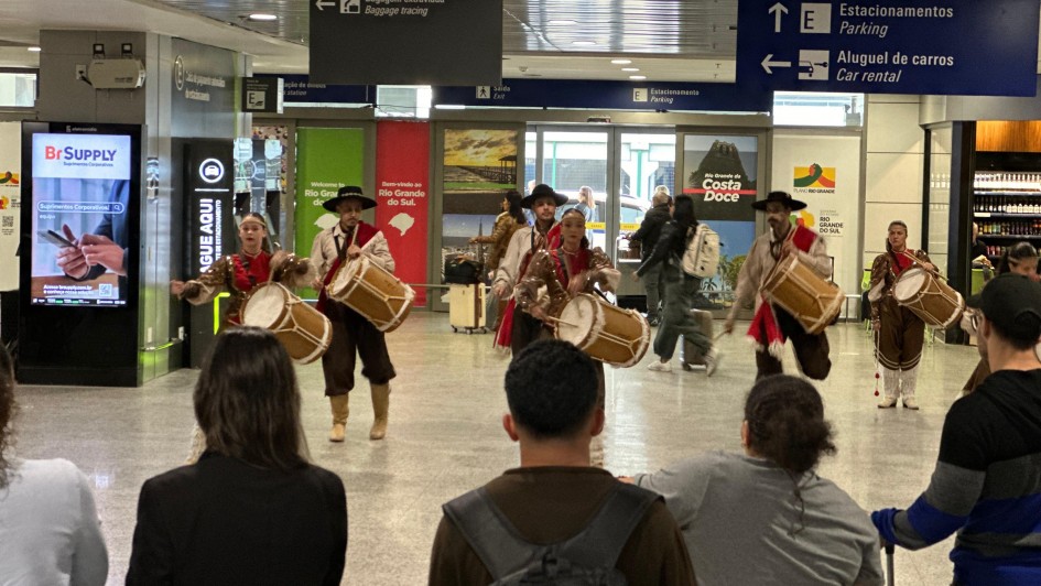 A imagem mostra um grupo de pessoas pilchadas tocando tambores em um sagu&atilde;o de aeroporto. Algumas pessoas se aglomeram ao redor para assistir a apresenta&ccedil;&atilde;o. 