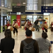 A imagem mostra um grupo de pessoas pilchadas tocando tambores em um sagu&atilde;o de aeroporto. Algumas pessoas se aglomeram ao redor para assistir a apresenta&ccedil;&atilde;o. 