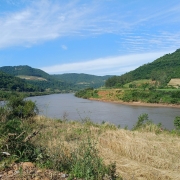 A fotografia apresenta uma vista panor&acirc;mica de um rio largo que serpenteia por um vale entre colinas cobertas de vegeta&ccedil;&atilde;o, sob um c&eacute;u azul claro.

Detalhes da Paisagem
O Rio: O curso d'&aacute;gua ocupa a parte central da imagem, apresentando &aacute;guas de tom acastanhado e uma superf&iacute;cie relativamente calma que reflete a claridade do dia.

O Relevo: Ao fundo e nas laterais, elevam-se morros e colinas cobertos por uma mata verde e densa. Algumas &aacute;reas nas encostas mostram clar&otilde;es de terra e campos cultivados, evidenciando o uso agr&iacute;cola da terra na regi&atilde;o.

Primeiro Plano: Na parte inferior da foto, v&ecirc;-se a vegeta&ccedil;&atilde;o da margem, composta por arbustos verdes, gram&iacute;neas secas de cor amarelada e um solo pedregoso no canto esquerdo.

O C&eacute;u: O c&eacute;u est&aacute; bem aberto e azul, com nuvens brancas finas e alongadas que se espalham de forma suave.

Contexto
De acordo com o t&iacute;tulo do arquivo, a imagem ilustra a implementa&ccedil;&atilde;o de um novo servi&ccedil;o de monitoramento pelo Governo do Estado do Rio Grande do Sul, projetado para prever o n&iacute;vel dos rios e auxiliar na preven&ccedil;&atilde;o de enchentes.