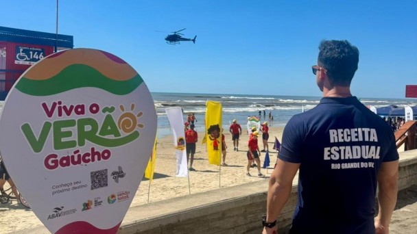 A imagem mostra um homem de uniforme da Receita Estadual de costas. Ao fundo, est&aacute; o mar e banhistas. H&aacute; uma placa no lado esquerdo onde se l&ecirc; "Viva o ver&atilde;o ga&uacute;cho". 