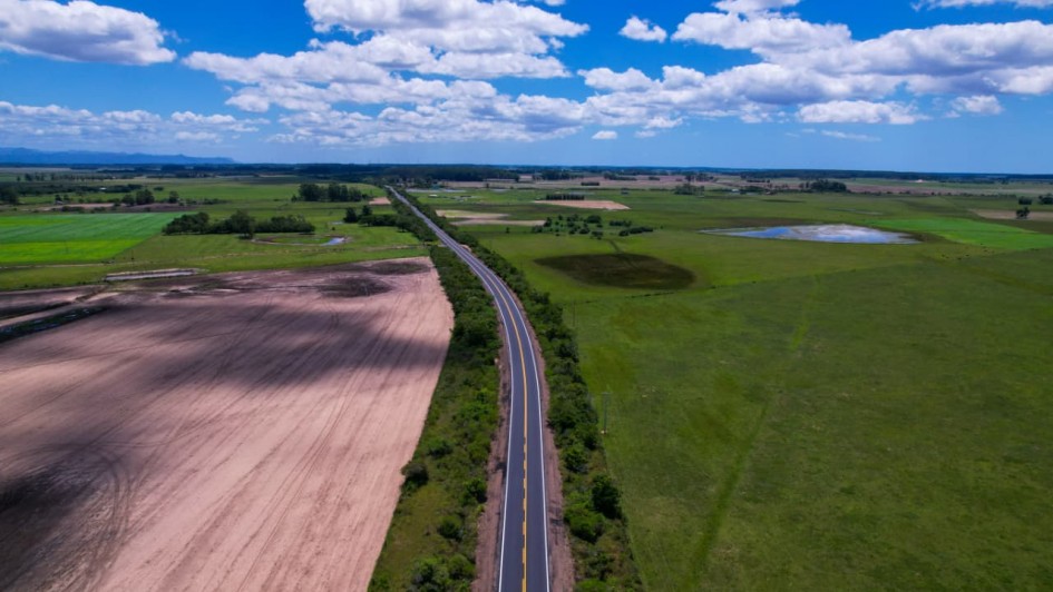 A fotografia a&eacute;rea, capturada de um &acirc;ngulo elevado (provavelmente por um drone), mostra um longo trecho retil&iacute;neo da rodovia RSC-101 atravessando uma vasta &aacute;rea rural sob um c&eacute;u azul vibrante.

Detalhes da Rodovia e Paisagem
A Estrada: O asfalto escuro da rodovia corta a imagem verticalmente, estendendo-se em linha reta at&eacute; o horizonte. A pista apresenta sinaliza&ccedil;&atilde;o nova, com faixas amarelas cont&iacute;nuas bem definidas no centro.

Vegeta&ccedil;&atilde;o: As margens da estrada s&atilde;o acompanhadas por uma linha de arbustos e vegeta&ccedil;&atilde;o rasteira verde. Ao redor, a paisagem &eacute; composta por um mosaico de grandes campos verdes e &aacute;reas de solo preparado para o plantio em tons de bege e marrom claro.

Elementos H&iacute;dricos: &Agrave; direita da estrada, nota-se um pequeno lago ou a&ccedil;ude de formato irregular que reflete a cor azul do c&eacute;u.

O C&eacute;u: Ocupa a parte superior da fotografia, exibindo um azul intenso com diversas nuvens brancas (c&uacute;mulos) espalhadas, cujas sombras podem ser vistas projetadas sobre os campos abaixo.

Contexto
De acordo com o t&iacute;tulo do arquivo, a imagem registra os resultados das obras de recupera&ccedil;&atilde;o da RSC-101, cujo contrato foi ampliado em 2025 com um investimento adicional de R$ 51,8 milh&otilde;es para garantir a qualidade da infraestrutura.