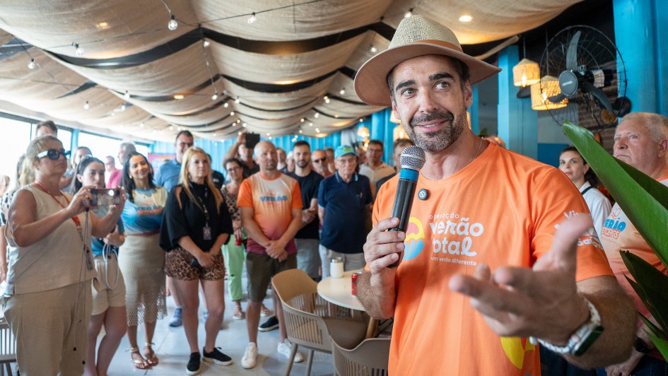 Homem com chep&eacute;u fala ao microfone para um p&uacute;blico formado por v&aacute;rias pessoas em p&eacute; em um ambiente coberto