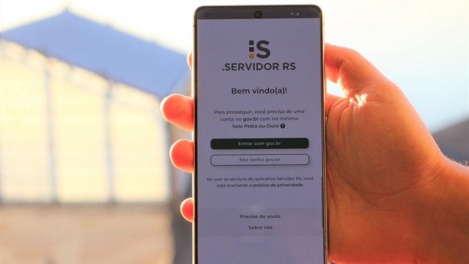 Foto mostra uma m&atilde;o segurando um celular e na tela a capa do aplicativo Servidor RS.