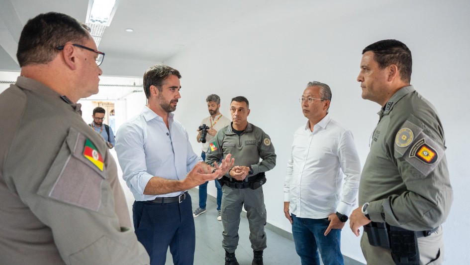Esta &eacute; uma descri&ccedil;&atilde;o da imagem para fins de acessibilidade:

Descri&ccedil;&atilde;o da Imagem

Uma fotografia colorida em plano m&eacute;dio mostra o governador do Rio Grande do Sul, Eduardo Leite, em uma conversa com oficiais da Brigada Militar e outros assessores em um ambiente interno.

Eduardo Leite: Localizado ao centro-esquerda da imagem, &eacute; um homem branco de cabelos castanhos curtos e barba aparada. Ele veste uma camisa social azul clara de mangas compridas (com as mangas levemente dobradas) e cal&ccedil;a social azul marinho. Ele est&aacute; de perfil para a direita, com as m&atilde;os erguidas e palmas voltadas para cima, em um gesto de explica&ccedil;&atilde;o ou di&aacute;logo.

Os Policiais: H&aacute; tr&ecirc;s oficiais da Brigada Militar vis&iacute;veis. No primeiro plano &agrave; esquerda, um oficial est&aacute; de costas, mostrando o detalhe da bandeira do Rio Grande do Sul e o bras&atilde;o da corpora&ccedil;&atilde;o no bra&ccedil;o do uniforme cinza-esverdeado. Ao centro, ao fundo, outro oficial observa a conversa. &Agrave; direita da imagem, um terceiro oficial est&aacute; de perfil, ouvindo o governador; ele possui um bras&atilde;o amarelo e vermelho no bra&ccedil;o direito.

Outras Pessoas: Entre Eduardo Leite e o oficial da direita, est&aacute; um homem de camisa social branca e cal&ccedil;a jeans, com &oacute;culos, mantendo uma postura de escuta. Ao fundo, no corredor, um fot&oacute;grafo com uma c&acirc;mera profissional pendurada no pesco&ccedil;o observa a cena, e outra pessoa caminha mais ao longe.

O Ambiente: O local &eacute; um corredor amplo e bem iluminado, com paredes brancas e lisas, piso cinza claro e lumin&aacute;rias de LED embutidas no teto. O aspecto &eacute; de um pr&eacute;dio governamental ou administrativo moderno.