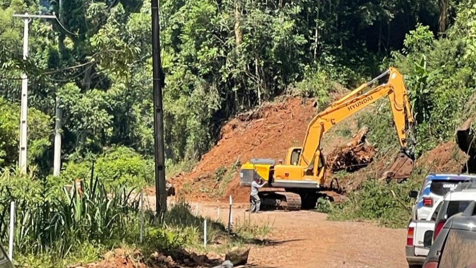 Foto de uma escavadeira sendo operada junto a morro, em &aacute;rea composto por uma mata ao fundo.