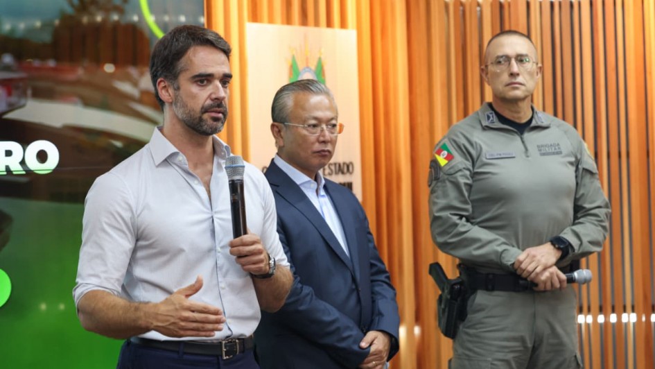 A imagem mostra o governador Eduardo Leite falando ao microfone. Ao seu lado, est&aacute; o titular da Seguran&ccedil;a P&uacute;blica e um policial militar fardado. 