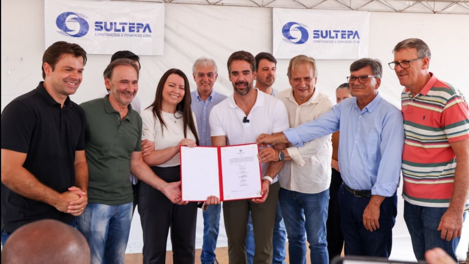 Um grupo de pessoas posa para a foto segurando um documento e sorrindo. Ao centro, est&aacute; o governador do RS