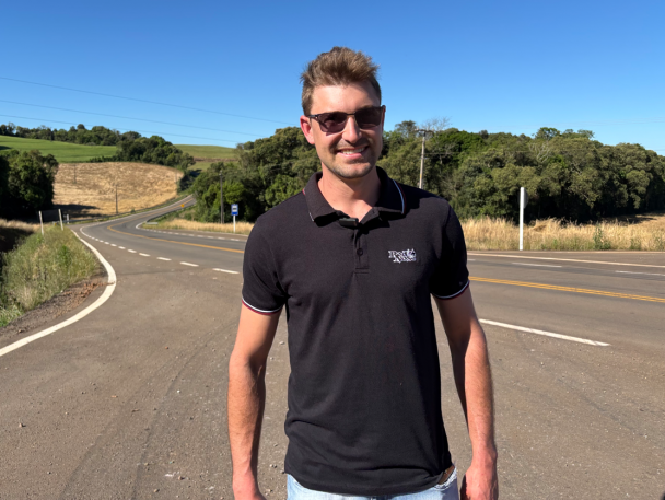 Foto mostra morador, com óculos escuros e camisa preta, sorrindo para a câmera. Ele está em frente à estrada. O céu está azul, sem nuvens. 