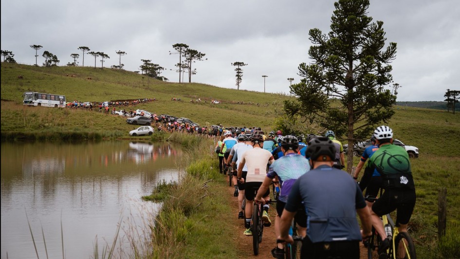 Foto em ambiente aberto, em meio a uma paisagem natural de grama, poucas arauc&aacute;rias e um riacho, no qual est&atilde;o reunidos, em fila, centenas de ciclistas pedalando. 