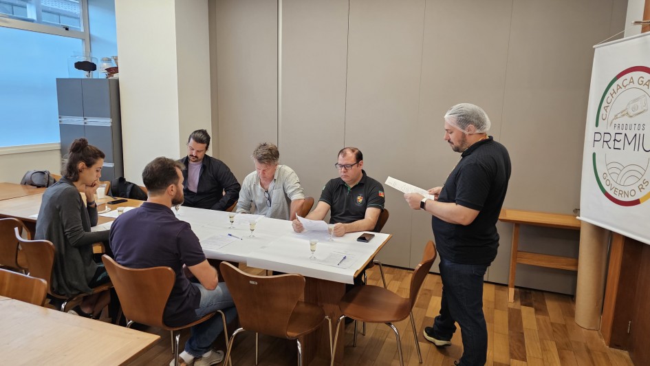Descri&ccedil;&atilde;o da Imagem
Uma fotografia colorida em plano m&eacute;dio captura seis pessoas em uma sala de reuni&otilde;es durante um processo de avalia&ccedil;&atilde;o t&eacute;cnica.

A&ccedil;&atilde;o Principal: Cinco pessoas (uma mulher e quatro homens) est&atilde;o sentadas ao redor de uma mesa retangular coberta com um papel branco que serve como toalha descart&aacute;vel. Eles est&atilde;o concentrados, analisando documentos e fichas de avalia&ccedil;&atilde;o. Sobre a mesa, al&eacute;m dos pap&eacute;is e canetas, h&aacute; pequenos copos de vidro (c&aacute;lices) com um l&iacute;quido transparente, sugerindo uma degusta&ccedil;&atilde;o t&eacute;cnica de cacha&ccedil;a.

O Coordenador: &Agrave; direita da imagem, um homem est&aacute; de p&eacute;, vestindo uma camisa polo preta e cal&ccedil;a jeans. Ele usa uma touca descart&aacute;vel branca na cabe&ccedil;a (item de higiene) e segura uma folha de papel, parecendo orientar ou acompanhar o trabalho dos avaliadores.

Os Avaliadores:

Na ponta esquerda, uma mulher de cabelos escuros presos em um coque observa atentamente os documentos.

Ao longo da mesa, os outros homens vestem roupas casuais (camisas polo e camisas de bot&atilde;o) e mant&ecirc;m o olhar fixo no material de an&aacute;lise.

O Ambiente:

&Agrave; direita, h&aacute; um banner branco com um logotipo circular onde se l&ecirc;: "CACHA&Ccedil;A GA&Uacute;CHA - PRODUTOS PREMIUM - GOVERNO RS".

A sala possui piso de madeira clara, paredes de pain&eacute;is cinzas e uma ilumina&ccedil;&atilde;o clara vinda do teto e de uma janela ao fundo, &agrave; esquerda.