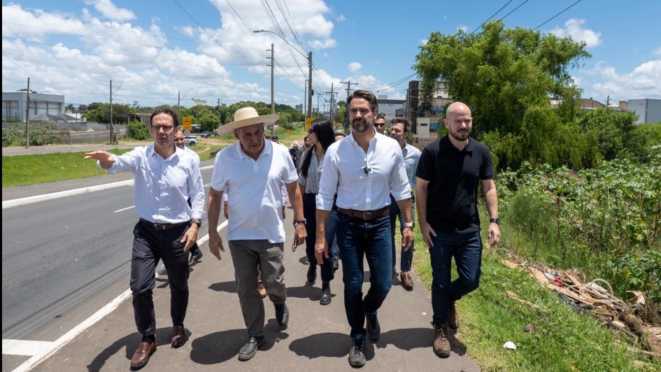 Um grupo de homens caminha por uma rua, ao ar livre, sob sol forte. Um dos homens aponta algo &agrave; frente, como se estivesse explicando o local. H&aacute; postes, vegeta&ccedil;&atilde;o e pr&eacute;dios ao fundo, e o c&eacute;u est&aacute; azul com nuvens.