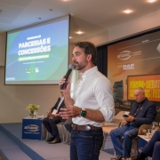 Uma fotografia colorida em plano m&eacute;dio captura o governador Eduardo Leite discursando durante um evento institucional em um audit&oacute;rio.

A&ccedil;&atilde;o Principal: O governador est&aacute; em p&eacute;, posicionado &agrave; direita do centro da imagem, falando ao p&uacute;blico. Ele segura um microfone preto com a m&atilde;o direita pr&oacute;xima ao rosto e gesticula com a m&atilde;o esquerda. Sua express&atilde;o &eacute; focada e comunicativa.

Eduardo Leite: Veste uma camisa social branca com as mangas dobradas, cal&ccedil;a jeans escura e um cinto de couro marrom. No pulso esquerdo, destaca-se um smartwatch com o visor digital aceso em verde.

O Cen&aacute;rio (Palco):

&Agrave; esquerda: Uma tela de proje&ccedil;&atilde;o exibe o t&iacute;tulo da apresenta&ccedil;&atilde;o: "PROGRAMA DE PARCERIAS E CONCESS&Otilde;ES - CONCESS&Otilde;ES DE RODOVIAS" sobre um fundo verde e azul.

Ao fundo: H&aacute; um painel (backdrop) com as logomarcas da SETCERGS, FEDERASUL, DAF e do Governo do Estado do Rio Grande do Sul. O painel tamb&eacute;m traz o t&iacute;tulo: "F&Oacute;RUM DE DEBATES SETCERGS | FEDERASUL".

Participantes: Dois homens est&atilde;o sentados em cadeiras beges atr&aacute;s do governador, ouvindo o discurso. &Agrave; direita, v&ecirc;-se parte de um p&uacute;lpito met&aacute;lico com o adesivo da SETCERGS.

O Ambiente: O audit&oacute;rio possui ilumina&ccedil;&atilde;o quente vinda de spots no teto, piso de madeira (parquet) e uma caixa de som branca montada em um trip&eacute; &agrave; esquerda.
