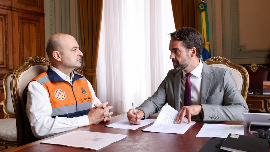 Dois homens est&atilde;o sentados em uma mesa de escrit&oacute;rio elegante, conversando e analisando documentos. Um veste terno e gravata, e o outro usa uniforme da Defesa Civil.