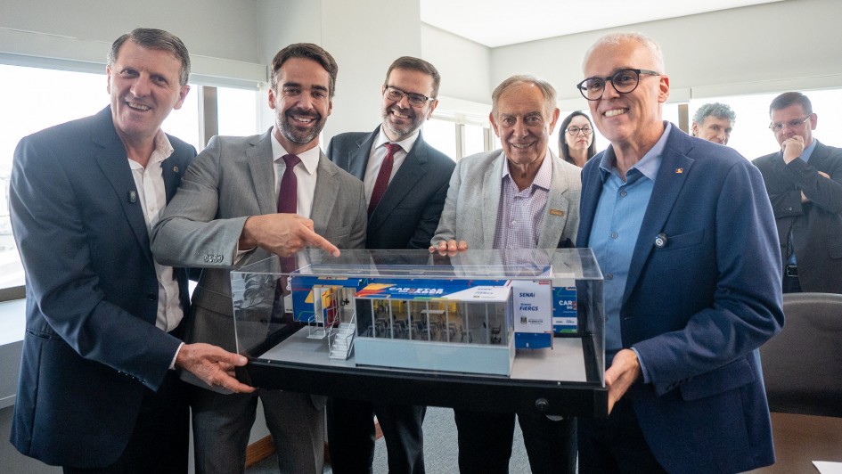 Uma fotografia colorida em plano m&eacute;dio captura o governador Eduardo Leite e outras quatro autoridades durante a assinatura de um conv&ecirc;nio em um gabinete.

A&ccedil;&atilde;o Principal: O grupo est&aacute; reunido em torno de uma maquete detalhada de uma unidade m&oacute;vel (carreta), protegida por um estojo de acr&iacute;lico transparente. Eles seguram a base da maquete e sorriem para a c&acirc;mera em sinal de celebra&ccedil;&atilde;o.

Eduardo Leite: O governador &eacute; o segundo da esquerda para a direita. Ele veste um terno cinza claro, camisa branca e uma gravata vinho. Com a m&atilde;o direita, ele aponta para a maquete, destacando o projeto.

Os Participantes: &Agrave; esquerda e &agrave; direita de Leite, quatro homens em trajes formais (ternos e palet&oacute;s em tons de azul e cinza) participam do registro. Ao fundo, &agrave; direita, veem-se outras duas pessoas observando a cena.

A Maquete: Representa uma unidade do programa "Carretas do Saber". &Eacute; um ve&iacute;culo azul e branco com &aacute;reas envidra&ccedil;adas que mostram um interior mobiliado como uma sala de aula ou laborat&oacute;rio. &Eacute; poss&iacute;vel ler as logomarcas "SENAI" e "FIERGS" na lateral do modelo.

O Ambiente: A sala &eacute; moderna e bem iluminada por janelas amplas ao fundo, que revelam uma vista externa clara. As paredes s&atilde;o de um tom cinza neutro, criando um aspecto profissional e s&oacute;brio.