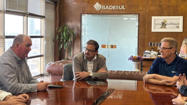 A imagem mostra tr&ecirc;s homens sentados em torno de uma mesa de reuni&atilde;o, conversando. Ao fundo, se l&ecirc; "Badesul". 