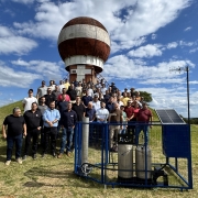 Foto posada ao ar livre de um grupo de mais de 50 pessoas de p&eacute;, em ambiente de campo aberto. Ao fundo h&aacute; uma estrutura redonda.