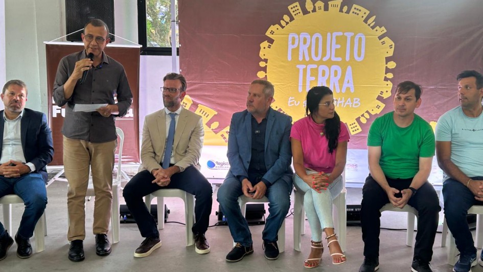 Esta &eacute; uma descri&ccedil;&atilde;o da imagem para fins de acessibilidade:

Descri&ccedil;&atilde;o da Imagem
Uma fotografia colorida em plano m&eacute;dio registra um evento institucional do governo para a entrega de escrituras a mutu&aacute;rios da extinta Cohab, no &acirc;mbito do Projeto Terra.

A&ccedil;&atilde;o Principal: Sete pessoas est&atilde;o posicionadas &agrave; frente de um banner informativo. Um homem &agrave; esquerda, vestindo uma camisa social marrom e cal&ccedil;a bege, est&aacute; em p&eacute; falando ao microfone enquanto consulta um papel em sua m&atilde;o.

Os Participantes: As outras seis pessoas (cinco homens e uma mulher) est&atilde;o sentadas em cadeiras brancas de pl&aacute;stico, ouvindo o discurso. Eles vestem trajes que variam do formal ao casual:

&Agrave; esquerda do orador, um homem de terno azul escuro.

&Agrave; direita do orador, um homem de palet&oacute; bege e gravata azul claro, seguido por um homem de blazer azul e uma mulher de camiseta rosa e cal&ccedil;a clara.

Na ponta direita, dois homens vestindo camisetas vibrantes (um verde e outro azul claro).

O Banner: Atr&aacute;s do grupo, h&aacute; um painel de fundo marrom com um grande sol amarelo centralizado. Dentro dele, est&aacute; escrito "PROJETO TERRA" e, logo abaixo, a frase "Eu sou COHAB". O desenho do sol &eacute; contornado por pequenas silhuetas de casas e pr&eacute;dios, simbolizando a regulariza&ccedil;&atilde;o fundi&aacute;ria.

O Ambiente: O local parece ser um pavilh&atilde;o ou centro comunit&aacute;rio com piso de concreto cinza e paredes claras. &Agrave; esquerda, atr&aacute;s do orador, observa-se uma caixa de som preta sobre um pedestal e parte de outro banner.